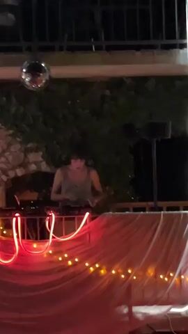 A person with short dark hair wearing a light-colored tank top stands behind a DJ setup. They are operating the equipment, with their hands visible on the controls. In the background, green laser lights are projecting, and a disco ball hangs from above. The DJ setup is draped with a light-colored fabric, adorned with red and yellow string lights. The environment appears to be outdoors, possibly a patio or balcony, with foliage and stone walls visible behind the DJ.