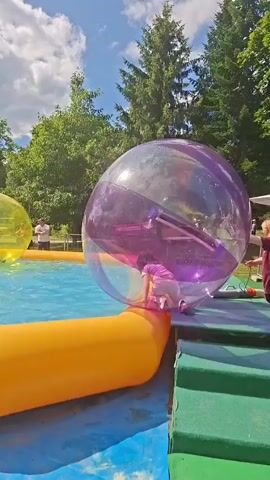 A person is inside a large, clear, purple inflatable sphere floating in a blue pool. The sphere is partially submerged in the water. Another yellow sphere is visible in the background to the left. Two people are standing near the edge of the pool, one on the right in a red shirt and another on the left in a white shirt. The pool is surrounded by an orange inflatable border. Green steps are visible on the right side of the pool, leading up to a wooden platform. Trees and a blue sky with white clouds are visible in the background. The person inside the sphere is a child wearing a pink and white outfit and appears to be moving around inside.