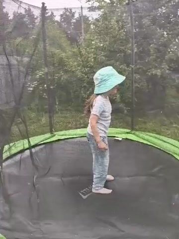 A girl in a light-colored hat, a striped t-shirt, and jeans jumps on a trampoline in a garden. The trampoline is black with a green rim, surrounded by a safety net. Trees and foliage are visible in the background under a cloudy sky. The girl jumps repeatedly, sometimes raising her arms, as the camera captures her from the waist up.