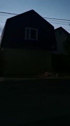 A dark, blurry video shows a residential area at dusk. The sky is a pale blue with hints of pink and orange. Two houses are visible, one dark and with a slanted roof, the other lighter with windows. Trees line the street, and power lines are strung overhead. A few cars are parked along the road, their shapes indistinct in the low light. The video appears to be shot from a moving vehicle.