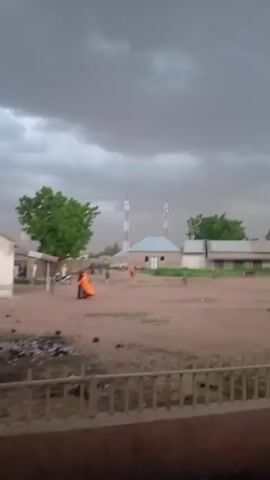 The video captures a scene in what appears to be a village under a cloudy, overcast sky, possibly indicating impending rain or a storm. In the background, several structures are visible, including houses with light-colored walls and roofs, and two tall towers that resemble communication masts. Green trees are scattered throughout the landscape. The ground appears to be dusty or made of reddish-brown soil. In the foreground, a person wearing a dark cap and a long, light-colored garment is seen walking away from the camera, towards the left. Another person, partially obscured, is seated to the left, wearing an orange garment over a dark fabric. The overall atmosphere is muted, with the sky dominating the frame with its dark clouds.