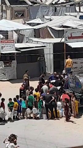 The video shows a crowd of people, including many children, gathered around a large tank or truck. They are holding white plastic containers, some of which are full of water, and appear to be waiting to collect water. The environment appears to be an outdoor setting, possibly a refugee camp, with tents and temporary structures visible in the background. The people are dressed in casual clothing and the overall atmosphere suggests a situation of scarcity or need, likely related to access to clean water.