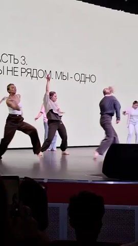 A group of dancers perform on a stage. The dancers are all wearing casual clothing, including pants and tops in various colors. They are barefoot. The backdrop of the stage is white with the text "ЧАСТЬ 3. МЫ НЕ РЯДОМ. МЫ - ОДНО" displayed. The dancers are performing a choreographed routine that involves various movements, including bending, reaching, and stretching. The camera captures different angles of the performance, and in the foreground, there are audience members watching. A speaker is partially obstructing the view of one of the dancers.