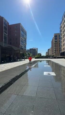 A young child wearing an orange jacket and grey pants is running in a paved outdoor area with a shallow water feature. The child's back is mostly to the camera as they run away from the viewer. The sun is shining brightly, casting a shadow of the child on the ground. Tall, modern apartment buildings are visible in the background, along with trees and a clear blue sky. There is a drain grate to the left of the child and square water jets are visible in the shallow water.
