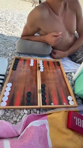 The video begins with a close-up of a backgammon board on a pebble beach. The board is set up for a game, with black and white checkers arranged in their starting positions. A man wearing sunglasses and a patterned swim trunk is lying on a towel behind the board, propped up on a grey pillow. The camera then pans slightly to the left, revealing a blue beach umbrella with white anchor patterns overhead. Beside the man, a red backpack and a mint green bag are placed on the towel. The background shows a crowded beach with people relaxing under umbrellas and sunbathing. The sea and a rocky cliff face are visible in the distance under a clear blue sky. The final shot is a wider view of the beach scene, emphasizing the sunny, vacation atmosphere.