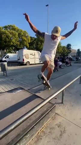 A man in a white tank top, tan shorts, and a tan baseball cap is skateboarding on a sunny day. He is performing a trick on a metal rail, with his body angled as he grinds on it. The skateboard is black with yellow wheels. In the background, there are trees, cars, buildings, and a van. The ground is paved with concrete and there is a ramp-like structure. The man has tattoos on his arms.