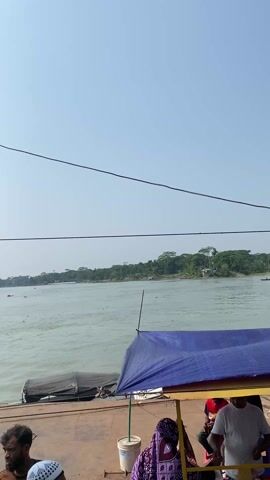 The video shows a wide river in a tropical landscape on a clear day. The view is taken from a ferry, with several people standing on the deck. A makeshift stall is visible in the foreground, covered by a blue tarp. The riverbank is lined with lush green trees and vegetation, including some flowering plants. A few small boats are docked or moving along the shoreline. A small, simple house is visible on the riverbank amidst the greenery. Powerlines run across the scene, slightly obscuring the view. The river appears to be somewhat murky, and there is some debris and vegetation floating in the water near the shore.