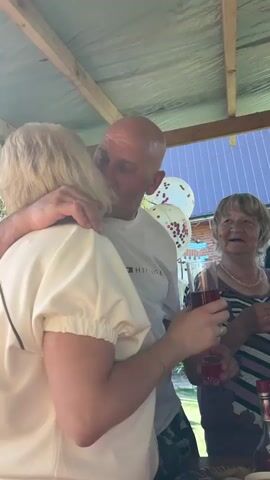 A group of people are gathered under a patio roof, celebrating and toasting with drinks. The video begins with a close-up of a man kissing a woman on the cheek. He is bald and wearing a white t-shirt, and she has blonde hair and is wearing a cream-colored top. Another older woman with blonde hair and a striped shirt is smiling in the background. The scene then expands to show more people around a table laden with food and bottles. The woman in the foreground holds up a glass of red liquid. Everyone appears to be in good spirits, smiling and laughing, as they raise their glasses for a toast. The outdoor setting is bright with sunlight.
