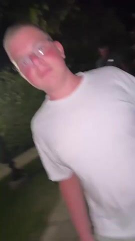 A person in a white t-shirt and light green shorts walks across a stone-paved area at night. The person is wearing glasses and has a red flush on their cheeks. They are carrying a white plastic bag in their right hand, which appears to contain food items. The background is dark, with some greenery and lights visible. Another person is partially visible in the background.