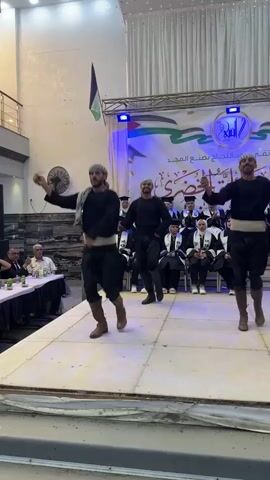 A group of men in traditional Palestinian clothing are performing a dance on a raised stage. They are wearing black vests, loose black pants, and headscarves. Two of the dancers are holding sticks and swinging them. In the background, there is a banner with Arabic writing and the Palestinian flag. Several people in graduation gowns are seated behind the dancers. The environment appears to be an indoor event space with white curtains and lights.