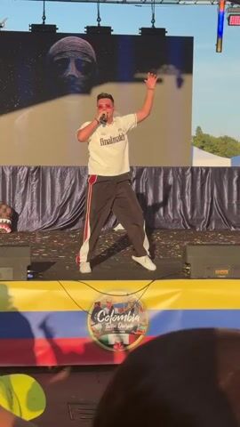A male performer, wearing a white shirt with red and black trim, black pants with red stripes, and pink sunglasses, stands center stage, holding a microphone and gesturing with his right arm. He is singing and dancing. A large screen behind him displays an image of a face. The stage is decorated with confetti. The stage front is adorned with the Colombian flag colors and a circular logo with the text "Colombia: Tierra Querida". In the foreground, the tops of spectators' heads are visible. The scene appears to be an outdoor event.