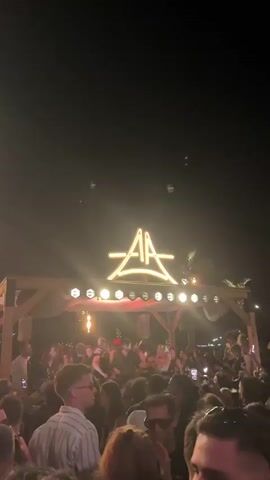 The video shows a crowded outdoor event at night. A large illuminated sign with the letters "AA" in a stylized font is visible above a structure with lights. A crowd of people is gathered below, many of whom appear to be dancing and holding up their phones, possibly recording the event. The lighting is bright and atmospheric, with some areas of fog or haze. The overall impression is that of a lively party or concert.