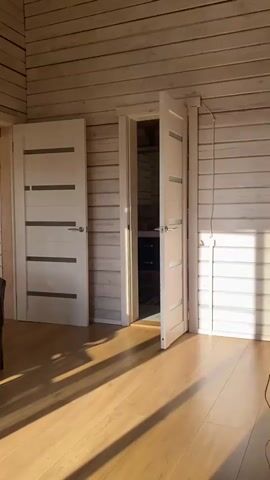 The video pans across a room with wooden plank walls and a wooden floor. There are several doors visible, some open and some closed. Sunlight streams in from the right, casting shadows on the floor and walls. In the foreground, a sofa is partially visible. Further into the room, a small kitchen area with cabinets, a microwave, and a stove is visible. Various electronic devices, including speakers and a laptop, are also present in the room. There are no people visible in the video.