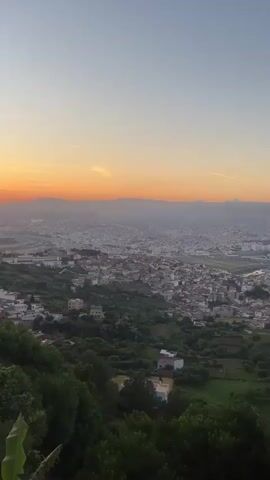 The image captures a panoramic view of the city of Tétouan, Morocco, at sunset from a high vantage point. The sky is a gradient of colors, with a vivid band of orange and yellow light along the horizon where the sun is setting, transitioning to a soft blue at the top. A faint, thin contrail is visible in the upper sky.

In the foreground, lush green vegetation, including trees and bushes, covers the hillside, with what appears to be banana leaves partially visible at the bottom left. A building with a reddish roof and light-colored walls is nestled among the greenery on the left side. To the far left, a rocky, lighter-colored hillside is visible, with a few parked cars and faint figures suggesting human presence.

The midground is dominated by the sprawling urban landscape of Tétouan, with numerous white and light-colored buildings densely packed across hills and valleys. Roads and other infrastructure are discernible within the city. The urban area extends towards the hazy distance, where distant mountains or hills are silhouetted against the bright horizon. The overall impression is one of a vast city enveloped by natural terrain, illuminated by the warm glow of the setting sun. No visible text or specific human activity can be clearly identified within the frame.