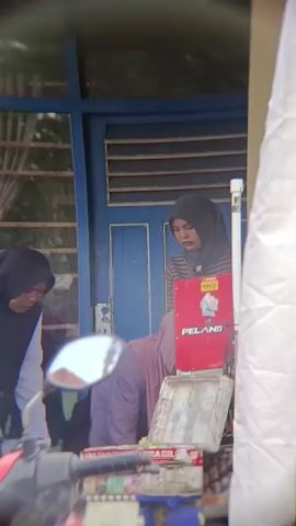A group of individuals are gathered outside a building. In the foreground, a red and black motorcycle is parked, with its handlebars and a portion of its body visible. Behind the motorcycle, several people are standing and interacting. Two individuals are wearing hijabs, one in pink and the other in black. Another person is wearing a white long-sleeved shirt and black pants, also wearing a black hijab. There is a red sign with the word "PELANGI" written on it, placed on a table or counter. In the background, the building appears to have blue doors and windows with blinds. The scene suggests an outdoor gathering or transaction taking place on a street or sidewalk.