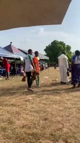 The video shows several people at an outdoor event, with a couple of them walking around holding live chickens. People are gathered under tents and in the open, some in formal attire and others in casual wear. One person is wearing a yellow shirt that reads "Our Lady of Mount Camel School". The ground is dry and covered in grass.