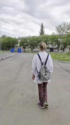 A young woman with blonde hair pulled back in a bun, wearing a white oversized shirt, camouflage pants, and a patterned backpack, walks away from the camera on a paved path. She turns to wave at the camera before continuing to walk away. The environment appears to be an outdoor urban or suburban setting with trees and buildings visible in the background. The sky is overcast.