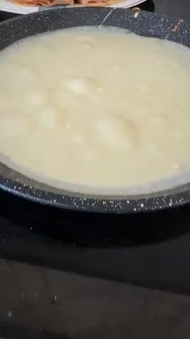 The video shows a close-up of a pan with a liquid batter for crepes. In the background, a plate of cooked crepes is visible, along with a spatula. The scene appears to be a kitchen, likely during the process of making crepes. The lighting is artificial, suggesting an indoor environment. The focus is on the food preparation, with the batter in the pan and the finished product in the background.