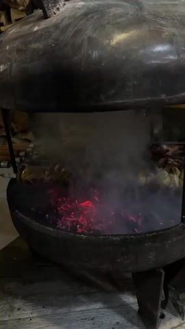 The video shows a close-up shot of a large, round, dark metal grill. Inside the grill, glowing red embers are visible, with smoke rising from them. Several skewers of food, appearing to be whole onions or potatoes, are arranged horizontally over the coals. The food is being cooked, and steam or smoke is billowing around it. The grill is supported by three dark metal legs, and the overall impression is one of an outdoor or rustic cooking setup. The lighting is dim, highlighting the intense heat of the embers and the cooking process.