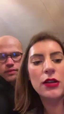 A video shows a man and a woman posing for the camera. The man is bald, wears glasses, and is in a black hoodie. The woman has brown hair, wears red lipstick, and is in a leather jacket. They are close together, and the woman is holding the camera. The background is blurry and indistinct, but appears to be indoors.