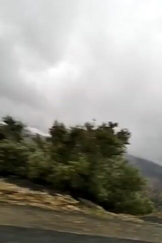 The video shows a view of a mountainous landscape under a heavily overcast sky. Patches of clouds cling to the mountain peaks in the distance, partially obscuring them. In the foreground, there are several green trees or bushes, and a paved road runs along the bottom of the frame. The camera appears to be moving, as the scenery shifts slightly. The overall tone of the video is gloomy due to the weather conditions.
