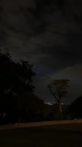 The video, recorded at night, depicts a grassy area bordered by trees and bushes. In the distance, a cityscape is visible, illuminated against a dark body of water. The sky is cloudy, with bright searchlights shining upwards, creating beams of light that cut through the night sky. The lights appear to be moving, with one beam flashing red at times. A faint voice is heard speaking in Spanish, expressing a desire to go towards the lights, but lamenting being unable to reach them.