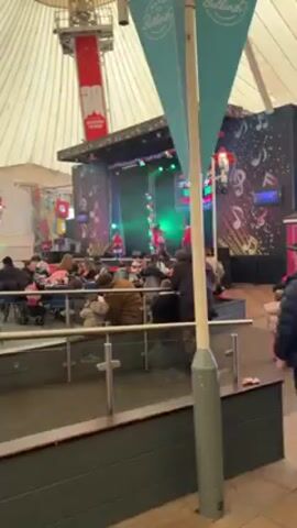 The video shows a stage performance with a group of performers in red outfits dancing on stage. The stage is decorated with lights and a backdrop featuring music notes. A crowd of people are seated at tables in front of the stage, watching the performance. Some people are standing and walking around. The setting appears to be an indoor event space with a large tent-like roof. A man in a black jacket and baseball cap walks through the crowd. There is a children's amusement ride visible in the background.