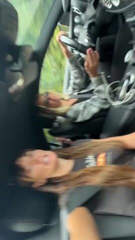 Two women are in a car. One woman is driving and wearing sunglasses and a plaid shirt. The other woman is in the passenger seat and is wearing a t-shirt with a croissant on it. The car is moving.