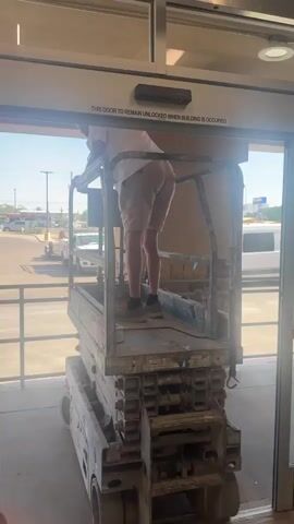 A person in a white t-shirt and khaki shorts stands on a blue scissor lift, working on a sliding glass door. The lift is inside what appears to be a commercial building entrance, with an automatic door sensor visible above the door. A ladder with a yellow cloth is placed to the left of the door. Outside the building, cars and a red truck are visible. The scissor lift has "GENIE" and "GS 3390" printed on its side. A sign on the door reads "THIS DOOR TO REMAIN UNLOCKED WHEN BUILDING IS OCCUPIED."
