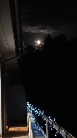 The video opens with a close-up shot of the moon in a dark, cloudy sky. The moon is bright and appears to be full. The camera then pans down to reveal a balcony decorated with string lights. In the background, a silhouette of trees can be seen against the night sky. The scene is dimly lit, with the only significant light sources being the moon and the string lights on the balcony. There are no people or animals visible in the video. The video appears to be a still shot with subtle movements in the clouds.