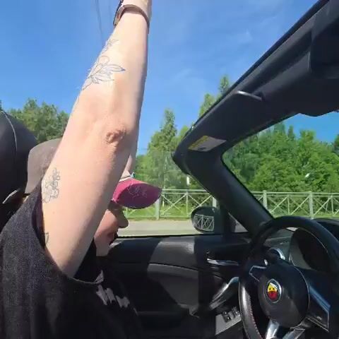 A person is driving a convertible car on a sunny day. The person is wearing a cap and has tattoos on their arm. The person raises their arm and waves. The top of the car is down, and the sky is blue with some clouds. The car is on a road with trees and a fence in the background.