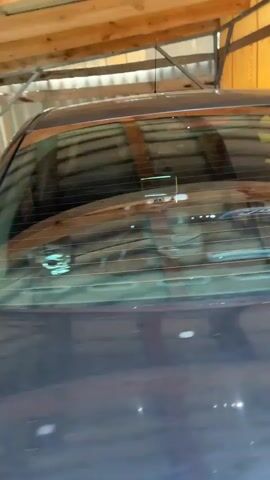 The video shows the interior and exterior of a dark-colored car parked inside a structure with corrugated metal walls and a wooden ceiling. The camera pans across the rear of the car, showing the rear window and part of the roof. It then moves to the side of the car, focusing on the rear door and window, and then the front door and window. The interior of the car is visible through the windows, including the steering wheel with a Ford logo, the dashboard, and the gear shifter. In the final shot, a man is seen in a reflection on the car window, holding a phone to take a selfie. He is shirtless and appears to be inside the car, looking at the camera.
