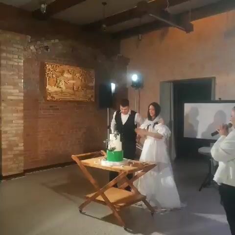 A wedding reception is underway in a dimly lit room with exposed brick walls and wooden beams. A bride in a white gown and a groom in a suit stand behind a wooden cake stand displaying a two-tiered cake with green frosting. The groom cuts the cake. A woman in a white jacket stands to the right, holding a microphone.