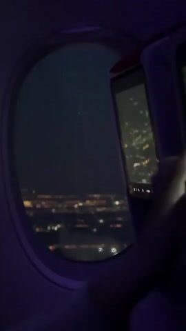 The video shows an airplane window at night, offering a view of a city illuminated by countless lights below. The external scene is blurred, creating a bokeh effect of bright, scattered lights against the dark sky. Inside the aircraft, the cabin has a dim, purplish ambient light. To the right of the window, a seatback entertainment screen is visible, displaying what appears to be a flight map or navigation interface with city outlines and light patterns. The overall impression is one of flying over a populated area at night, possibly Jianyang City, China, as suggested by the context.