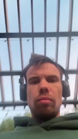 A first-person perspective video shows a man with dark, spiky hair and headphones around his head. He is wearing a green hoodie and looking directly at the camera with a serious expression. The background consists of a ceiling structure with horizontal slats and a light blue sky visible through the gaps. The scene appears to be outdoors, possibly on a patio or under a pergola. The man's head and shoulders are the primary focus, with the background slightly blurred, especially in the sections where trees and greenery are visible, indicating movement. The lighting is natural, suggesting it is daytime.