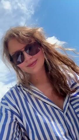 A woman with shoulder-length brown hair, wearing sunglasses and a blue and white striped shirt, smiles into the camera. She is outdoors near a body of water with a dirt path and green vegetation. The sky is blue with white clouds. There are cars parked near the water in the background. The woman is holding her phone at a slight downward angle, suggesting she is taking a selfie.