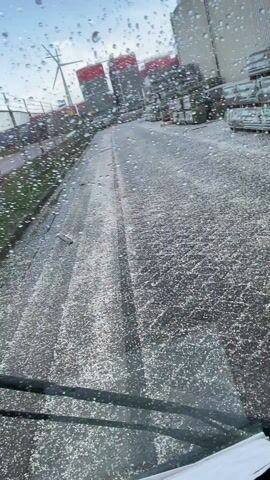 The video captures a hailstorm from the interior of a vehicle. The initial view is through the wet windshield, showing numerous small, white, round pellets (hail or sleet) impacting the glass and covering the dark, paved ground outside. In the background, large industrial buildings, some red and some light-colored, are visible, along with what appears to be stacked construction materials or pallets. A wind turbine can be briefly seen in the far left background.

The sound of the hailstones striking the vehicle's windows is prominent and continuous throughout the clip. Around the 0:02 mark, the camera briefly zooms in on the windshield, highlighting the water droplets and hailstones clinging to the glass. Shortly after, the windshield wipers activate, sweeping across the glass and clearing some of the precipitation, offering a momentary clearer view of the scene outside.

The camera then pans down and to the right, showing a side window of the vehicle. Through this window, the hail is visible covering a patch of green grass adjacent to the paved area, and a chain-link fence is also in view. The ground outside remains heavily speckled with the white hailstones, indicating a significant fall. The overall impression is of harsh, cold weather conditions.