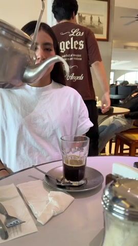 A woman is sitting at a table as a silver teapot pours milk into a glass filled with coffee. She's wearing a white T-shirt and looks straight ahead. The glass is on a small silver saucer with a spoon next to it. Two white napkins are on the table as well. A man wearing a brown "Los Angeles" T-shirt walks behind her.  There are other people in the background, sitting at tables. The setting appears to be a cafe or restaurant.