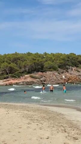 The video shows a beach with calm water and a wooded hill in the background. Several people are visible in the water, some of whom appear to be playing a game, possibly with a ball. The water is clear and a light blue, with small waves lapping at the shore. The sand on the beach is light brown and appears to be a typical sandy beach. The sky is blue with some scattered clouds. The overall atmosphere appears to be one of leisure and recreation on a sunny day.