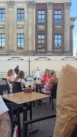 A person in a white wig and a white dress stands in front of a food truck with "Croissanterie" written on it. The person is holding a red object to their mouth. There are several people sitting at tables in front of the food truck. The environment appears to be an outdoor cafe or dining area. The building behind the food truck has classical architectural features.