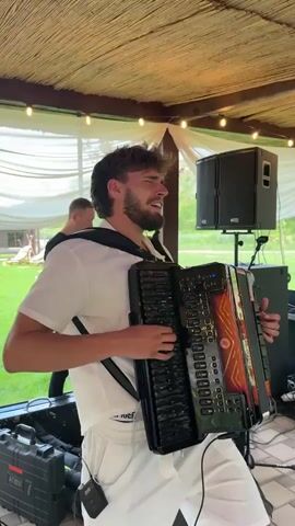 A young man with dark, curly hair and a beard is playing an accordion. He is wearing a white shirt and white shorts. He has earbuds in his ears and appears to be singing. The accordion is black with orange accents and has the brand name "Bugari EVO" on it. He is standing outdoors under a thatched roof structure with lights strung across it. In the background, another man is standing and looking at his phone. There are speakers and equipment cases near the accordion player. The ground appears to be paved with stone tiles.
