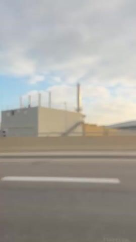 The video shows a view from a vehicle moving along a road. In the foreground, there is a road surface with a white line. A concrete barrier with alternating red and white stripes separates the road from the area beyond. In the background, there are several industrial or airport-related buildings. These buildings are light-colored and appear to be modern in design. Some have visible pipes or chimneys. The sky is partly cloudy with patches of blue sky visible. The lighting suggests it is daytime, possibly morning or late afternoon given the angle of the sun casting shadows. The overall impression is of an urban or industrial landscape seen from a moving vehicle.