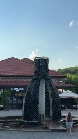 The video shows a large, decorative fountain shaped like a champagne bottle. The fountain is located in an outdoor plaza in front of a building with a red tiled roof and the words "CHAMPAGNE BAR" displayed prominently on its facade. A young girl with long brown hair and wearing a denim skirt and a brown top stands to the right of the fountain, facing away from the camera. She appears to be interacting with the fountain, possibly playing or posing. The fountain itself is a dark, cage-like structure adorned with what appear to be numerous small bottles, creating a lattice effect. A central column within the bottle shape appears to be the source of the water jets that shoot upwards and dissipate into a mist. The scene is set during the daytime, under a bright blue sky with scattered clouds. Trees and greenery are visible in the background, suggesting a pleasant, possibly resort-like environment. The overall atmosphere seems relaxed and cheerful. The visible text "CHAMPAGNE BAR" clearly indicates the nature of the establishment.