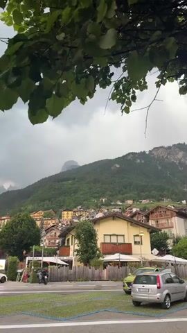 This vertical video captures a scenic view of Molveno, Italy, likely during a cloudy or overcast day. The foreground features a parking lot with several cars, including a silver SUV and a white station wagon. Beyond the parking lot, a row of buildings is visible, many with traditional alpine architecture, featuring wooden balconies and pitched roofs. One building has a sign that reads "GELATERIA" and another with "SNACK & DRINK". A motorcycle is parked in front of one of these establishments.

The middle ground is dominated by a hillside town, with colorful houses clustered together and ascending the green slopes of a large mountain. The mountain itself is covered in dense green forest, with rocky peaks visible in the distance. The sky is heavy with clouds, suggesting an impending rain shower or a generally overcast condition. The overall mood is serene and picturesque, characteristic of a mountain town setting. The presence of cars and the signs for a gelateria and snack bar indicate that this is a public area, likely a street or plaza in the town. The time of day appears to be daytime, but the cloudy weather obscures direct sunlight.