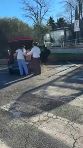 Two people are unloading items from the open trunk of a dark-colored car in a parking lot. One person is wearing light blue pants, while the other wears dark pants. A bus shelter is visible nearby. The camera then pans over to show other parked cars, including a silver car. In the background, a building sits atop a hill against a bright blue sky. The parking lot has cracked asphalt with painted parking lines. Shadows from trees are cast across the lot.