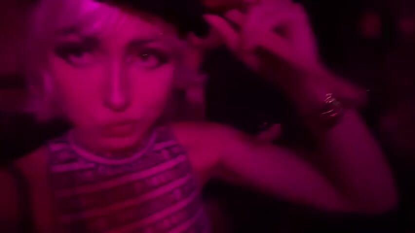 A person with short blonde hair, wearing a black hat, heavy eye makeup, and a black and white patterned top is shown in the foreground, illuminated by pink lighting. The person is making a kissing face and touching their hair. In the background, out of focus, another person is visible, also illuminated by pink lighting. The environment appears to be a dimly lit indoor space, possibly a club or party.