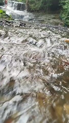A fast-flowing stream is shown from a low angle, with the water dominating the foreground. In the background, a small waterfall cascades down rocks, with trees and foliage lining the banks. A person in red shorts and a white top is visible standing on the rocks near the top of the waterfall. The water appears clear and is creating ripples and white foam as it moves.