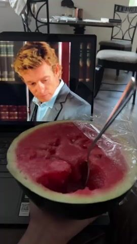 A person holds a half of a watermelon in their left hand, with a fork sticking out of it. The watermelon is positioned in front of an open laptop, displaying a scene from a television show or movie. The laptop is on a dark-colored keyboard. In the background, a window with light filtering through is visible, along with furniture and a chair. The footage transitions through several scenes on the laptop screen, including a close-up of a dark stain on a carpet, a man in a suit in an office, a close-up of a man's face, and a pile of papers with a calculator on top.