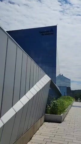 The video captures an outdoor scene on a cloudy day, likely a university campus, with modern architecture. The camera pans to the left, revealing a tall, dark blue glass building with "Université de Montréal" written in white on its upper facade. In the foreground, a grey, angular wall structure with diagonal metal panels runs along a paved walkway. Green foliage grows in planters between the walkway and the building. The walkway itself is made of light-colored rectangular paving stones. Beyond the initial building, more glass-faced structures are visible, as well as trees and hills in the distance. The sky is filled with scattered white clouds against a pale blue background. The camera then pans right, showing a wider view of the campus with multiple glass buildings and the paved walkway extending into the distance. The scene appears peaceful and quiet, with no people visible.