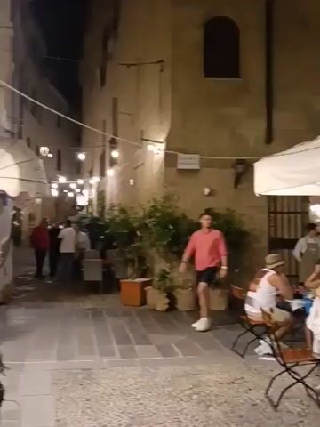 The video shows an outdoor dining area at night. In the foreground, a man in a red shirt and blue shorts walks past several tables where people are seated. There are umbrellas providing shade over some of the tables. The background shows buildings with lights on, and some of the buildings appear to be shops or restaurants. The overall atmosphere is that of a lively evening in a town or city.